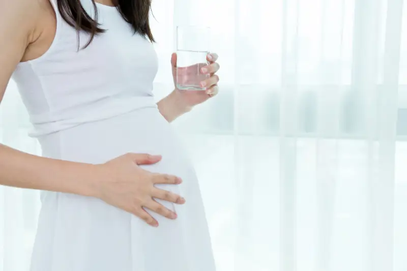 Close-up of a pregnant woman wearing a white dress, gently holding her belly with one hand and a glass of water in the other, standing by a window with sheer curtains.