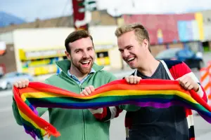 Two joyful men, holding a rainbow pride scarf, smiling outdoors. This represents the hope and inclusivity for gay and HIV-positive men pursuing fatherhood through IVF and surrogacy.
