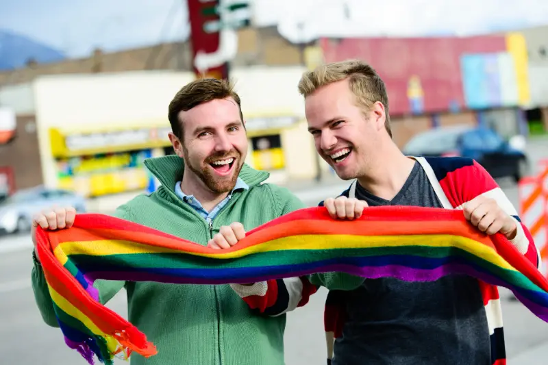 Two joyful men, holding a rainbow pride scarf, smiling outdoors. This represents the hope and inclusivity for gay and HIV-positive men pursuing fatherhood through IVF and surrogacy.