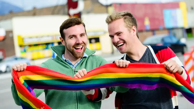 Two joyful men, holding a rainbow pride scarf, smiling outdoors. This represents the hope and inclusivity for gay and HIV-positive men pursuing fatherhood through IVF and surrogacy.
